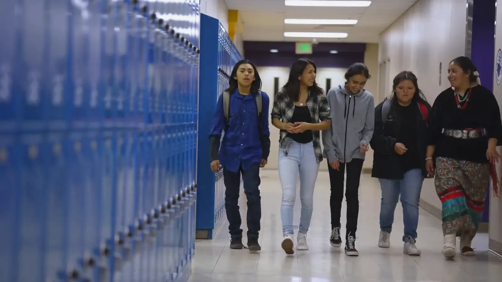 A group of students walks and talks together down a school hallway lined with blue lockers.