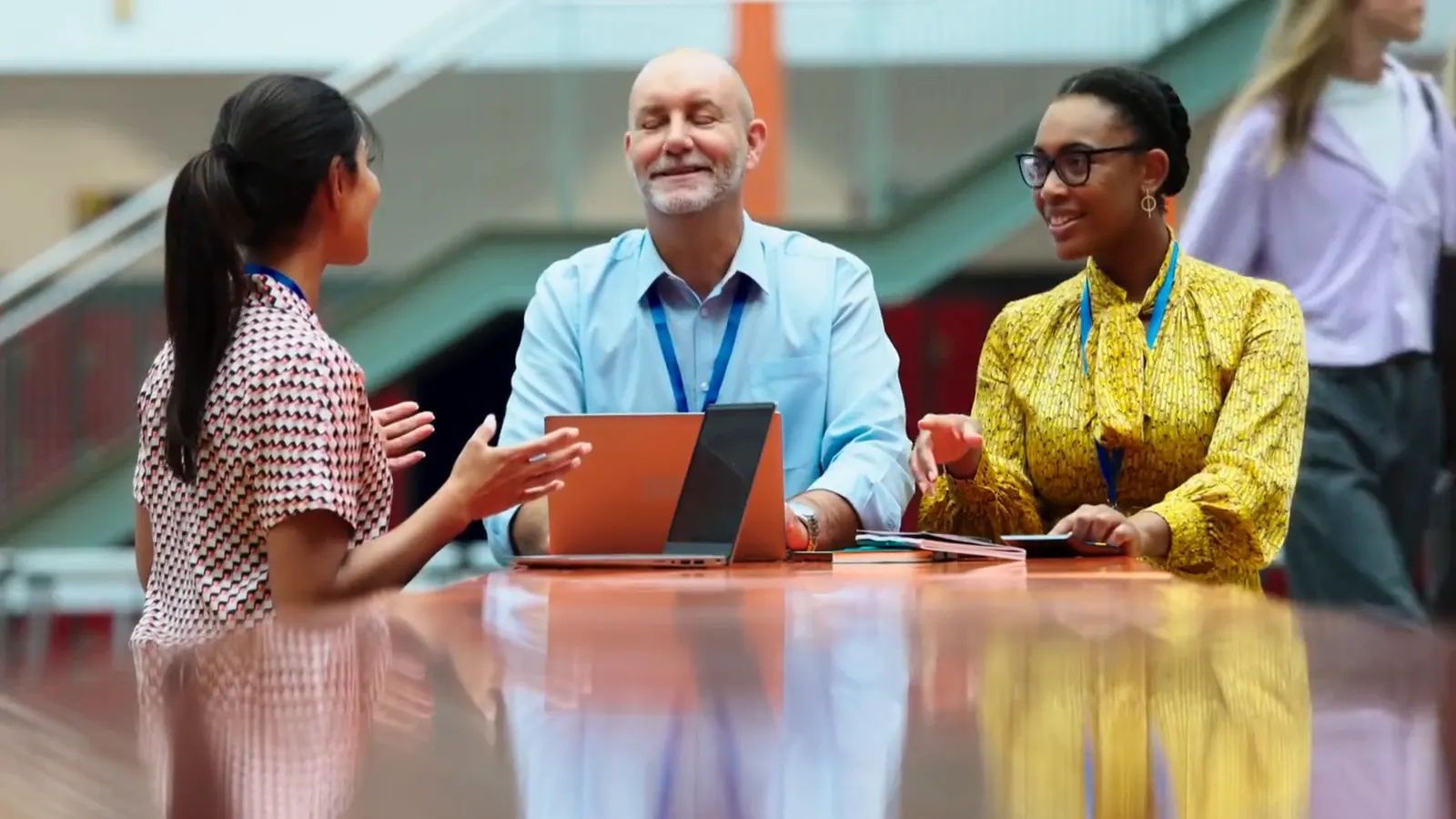 Two teachers talk and laugh with a student at a table in a busy school atrium.